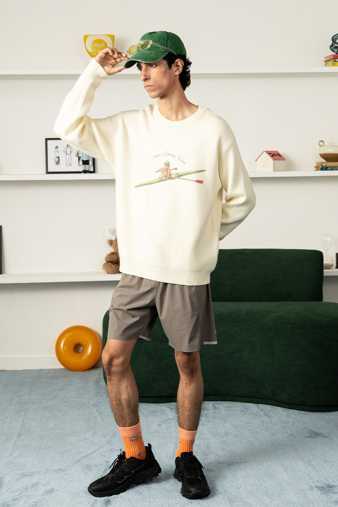 A man wearing a cream crewneck sweatshirt with a rowing graphic, gray shorts, orange socks, and black sneakers, standing in a modern room with a green couch and white shelves.