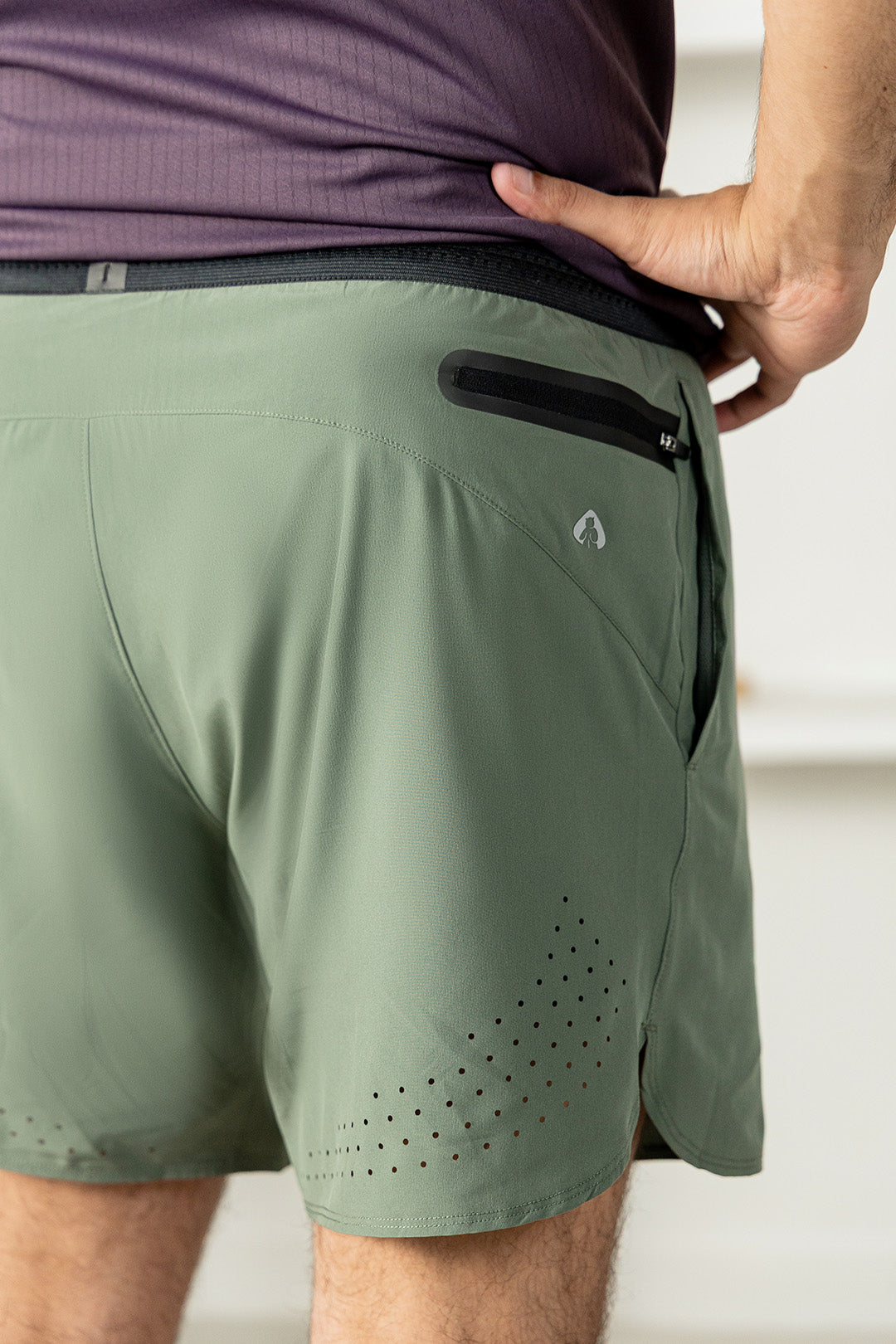 Image of Close-up of a person wearing olive green athletic shorts, showing the back pocket with a black zippered compartment and a small white logo, paired with a purple shirt.