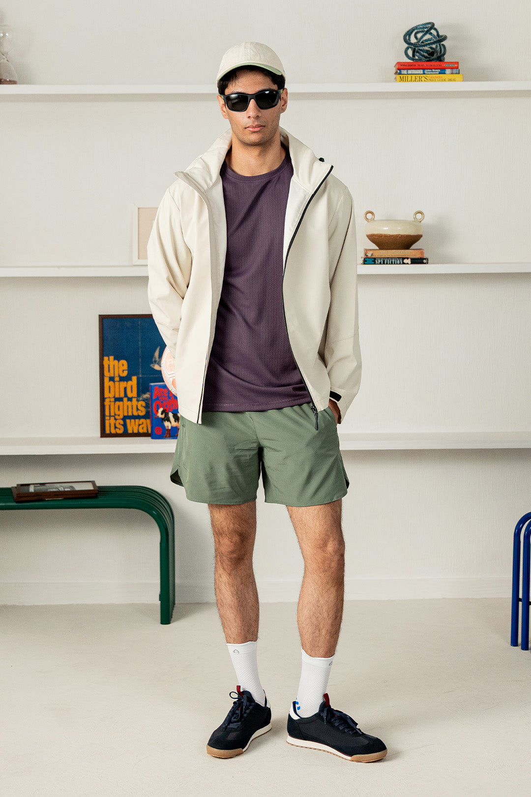A man modeling a light beige lightweight zip-up jacket over a dark purple t-shirt, olive green shorts, white socks, and navy sneakers, standing in a bright room with white shelves in the background.
