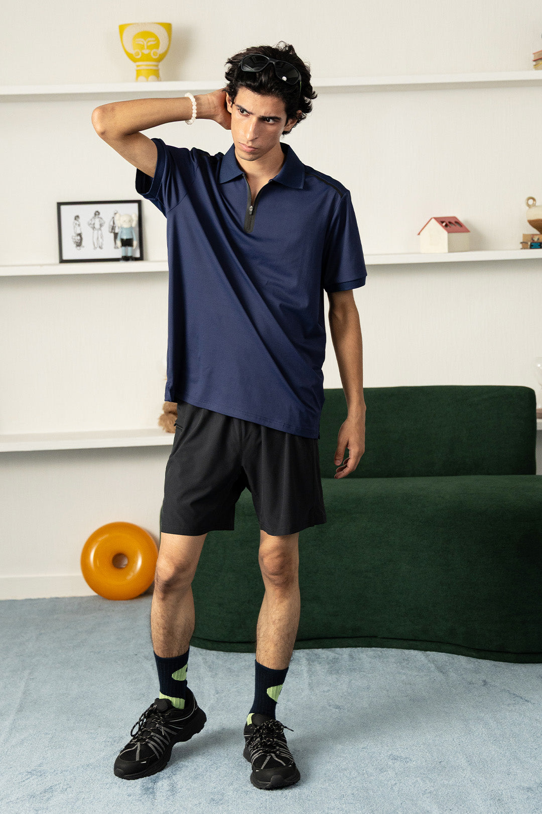 Image of A man modeling a navy blue short-sleeve polo shirt and black athletic shorts in a modern living room setting with a green sofa and light blue carpet.