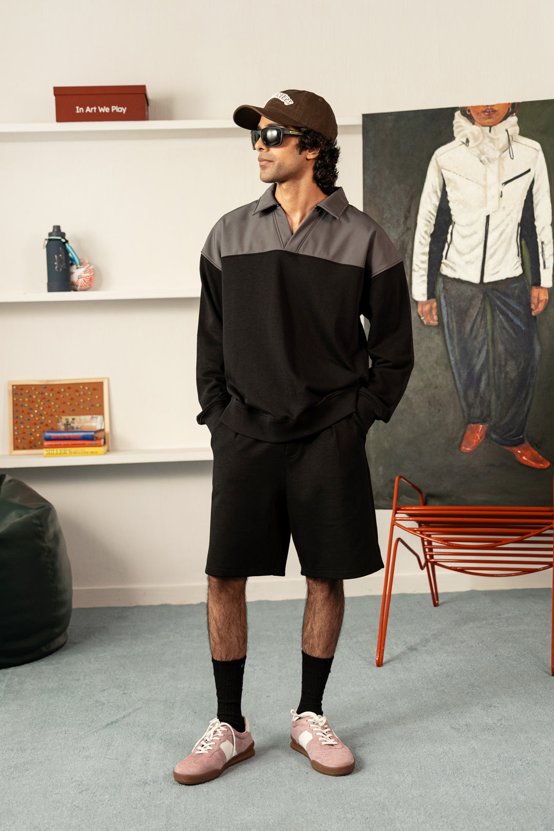 A man wearing a black and gray color-blocked long-sleeve polo shirt, black shorts, black socks, pink sneakers, and a brown baseball cap, standing in a styled room with shelves, a painting, and a red chair.
