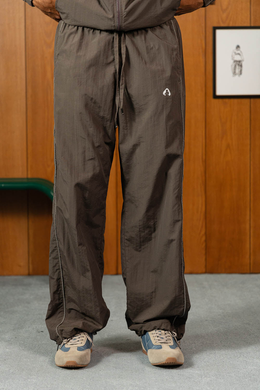 A person wearing dark brown wide-leg pants with a small white logo on the upper left thigh, paired with beige and blue sneakers, standing in front of a wood-paneled wall.