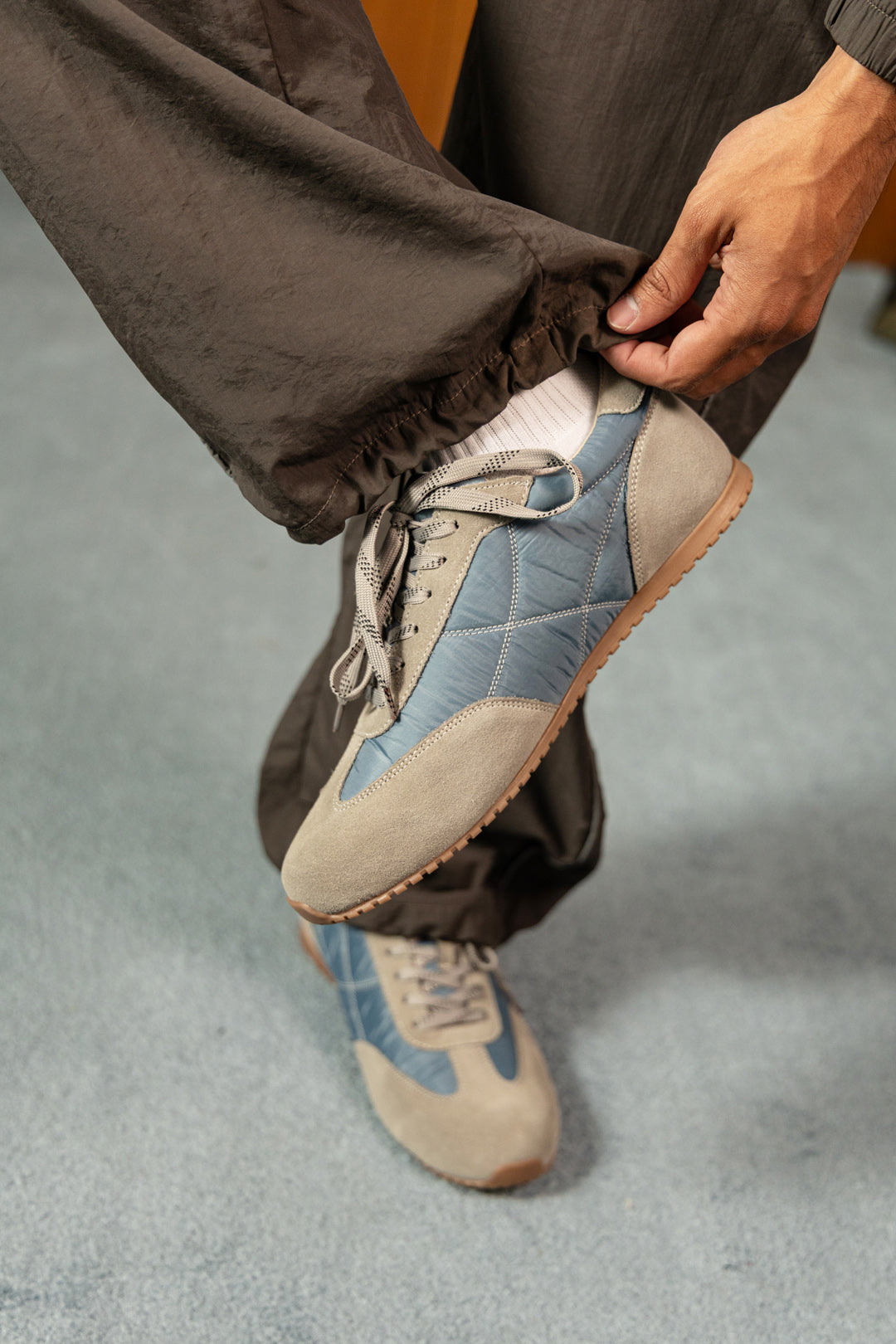 Image of A person wearing loose dark brown pants and light blue and beige sneakers, adjusting the cuff of the pants with one hand while wearing the shoes.