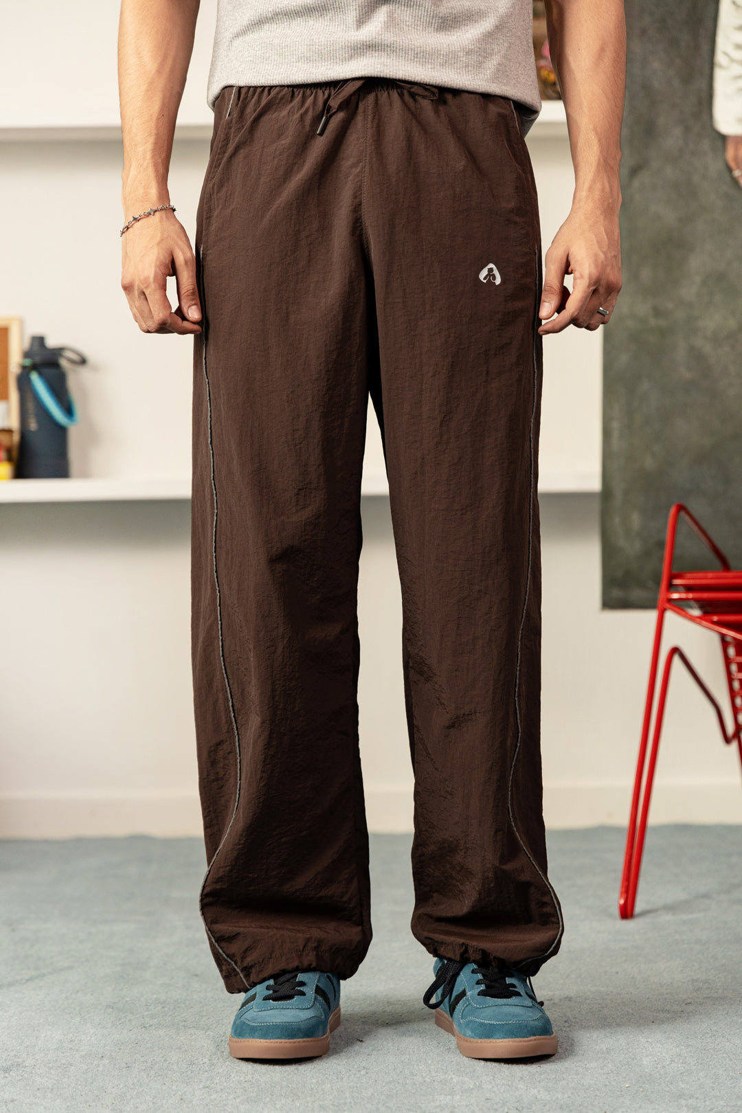 Image of A person wearing loose brown drawstring pants with a small white logo on the upper right thigh, paired with a light gray t-shirt and blue sneakers, standing in an indoor room.