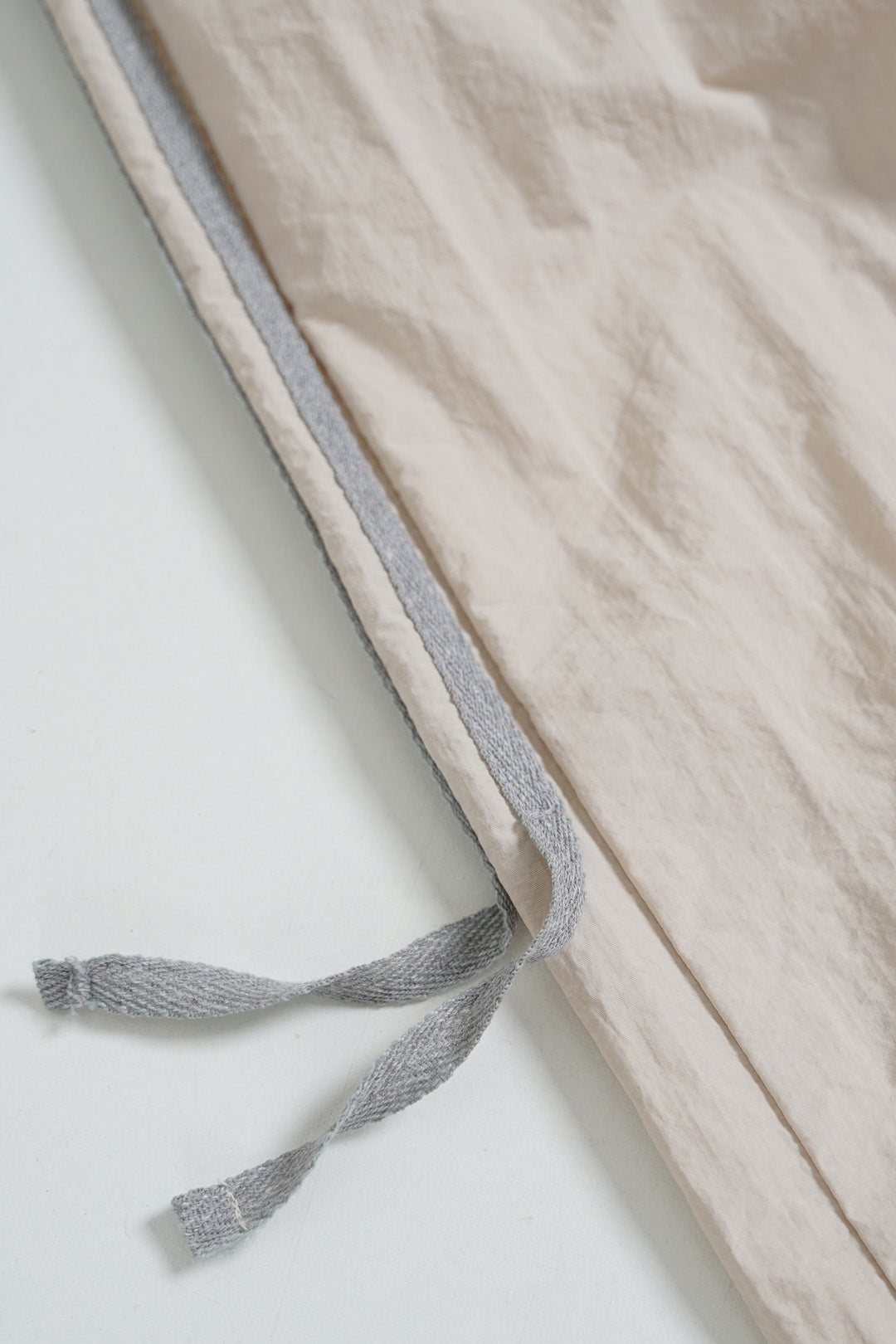 Image of A close-up of a light beige textured fabric item, likely a blanket or quilt, with a contrasting gray stitched border and matching gray drawstring ties, laid flat on a white surface.