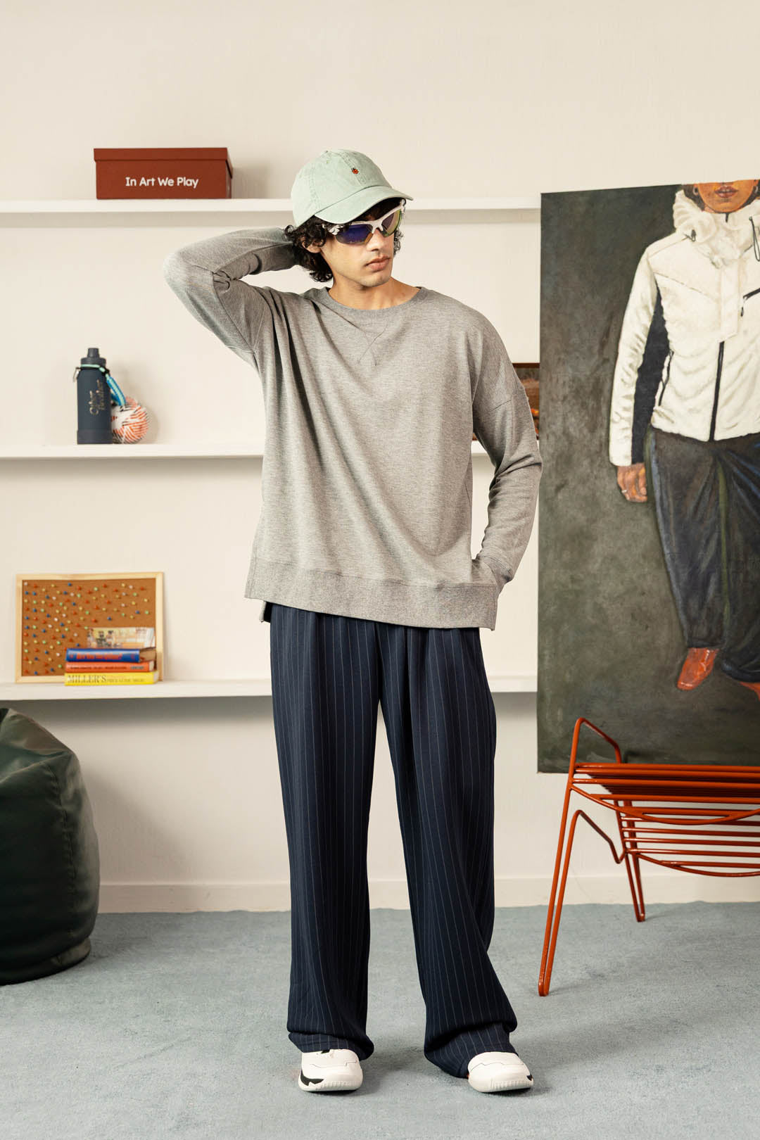 A person stands in a styled indoor room wearing an oversized light gray sweatshirt, dark navy pleated trousers, a pale green cap, sunglasses, and white sneakers. White shelves, a large portrait painting, and an orange chair are visible in the background.