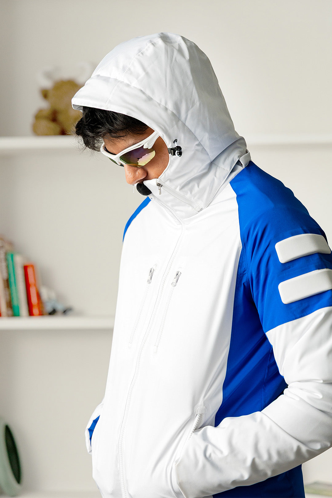 Image of A man wearing a white and blue ski jacket with a white hood, white ski goggles, and looking down, set against a bright indoor background with white shelves.