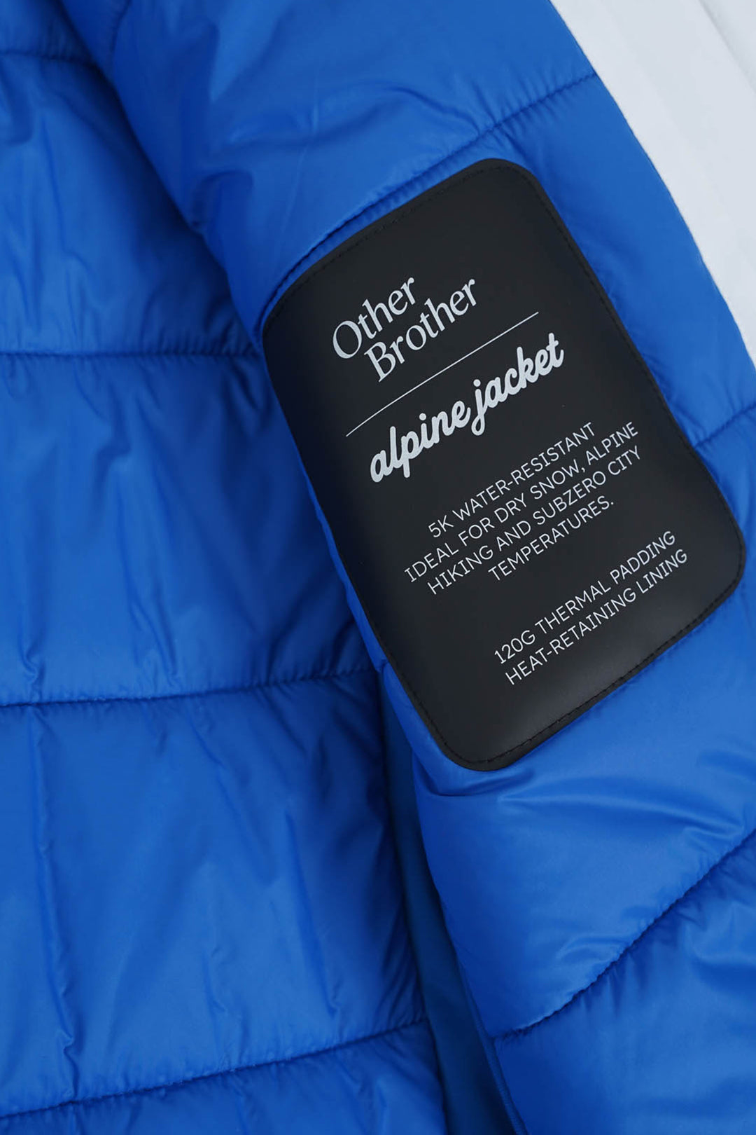 Image of Close-up of a bright blue puffer jacket with a black sewn-on brand patch reading 'Other Brother alpine jacket' on the chest.