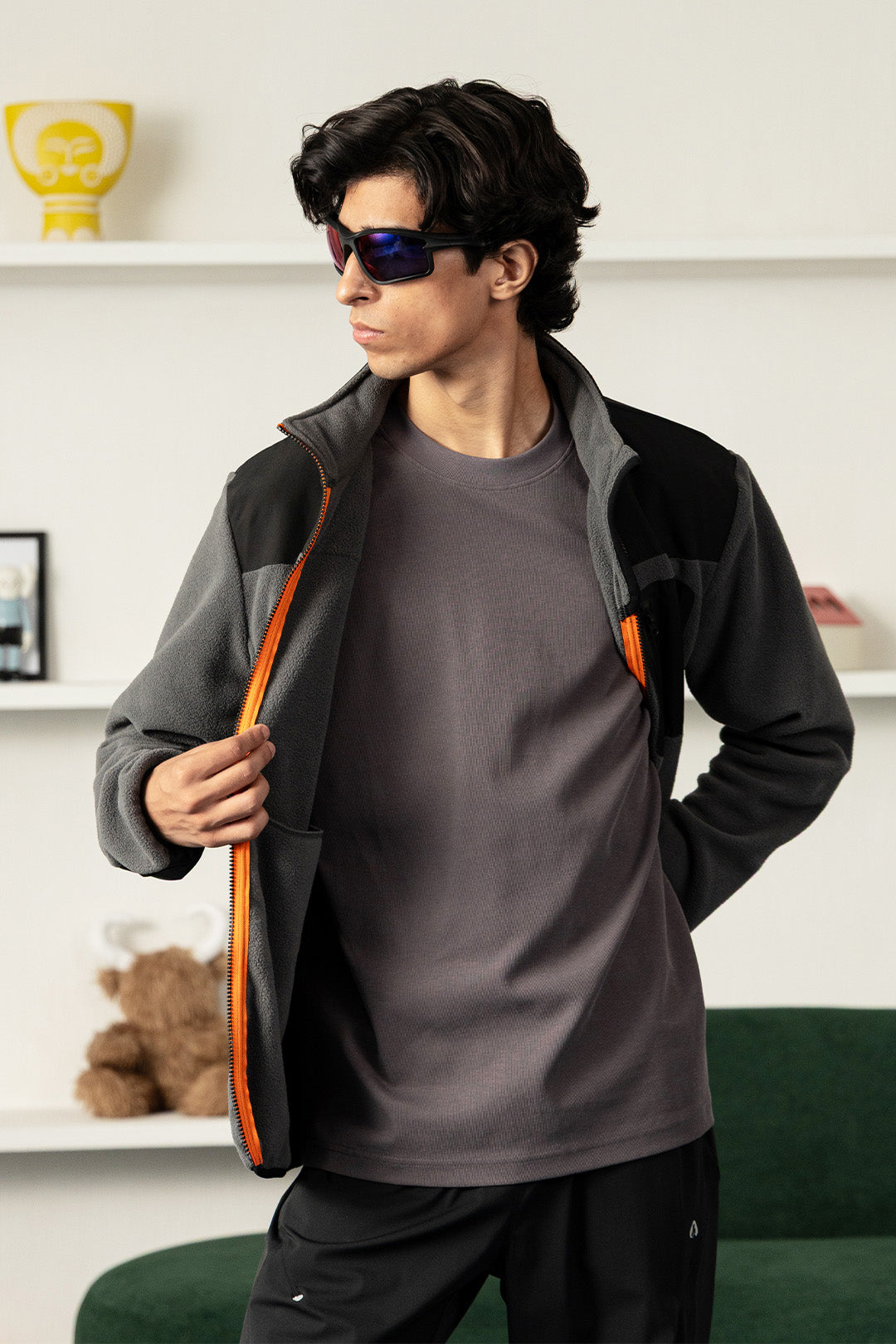 Image of A man wearing a dark gray and black zip-up fleece jacket with bright orange zipper accents, a dark gray t-shirt, and black pants, standing in a modern room with white shelves and a green sofa.