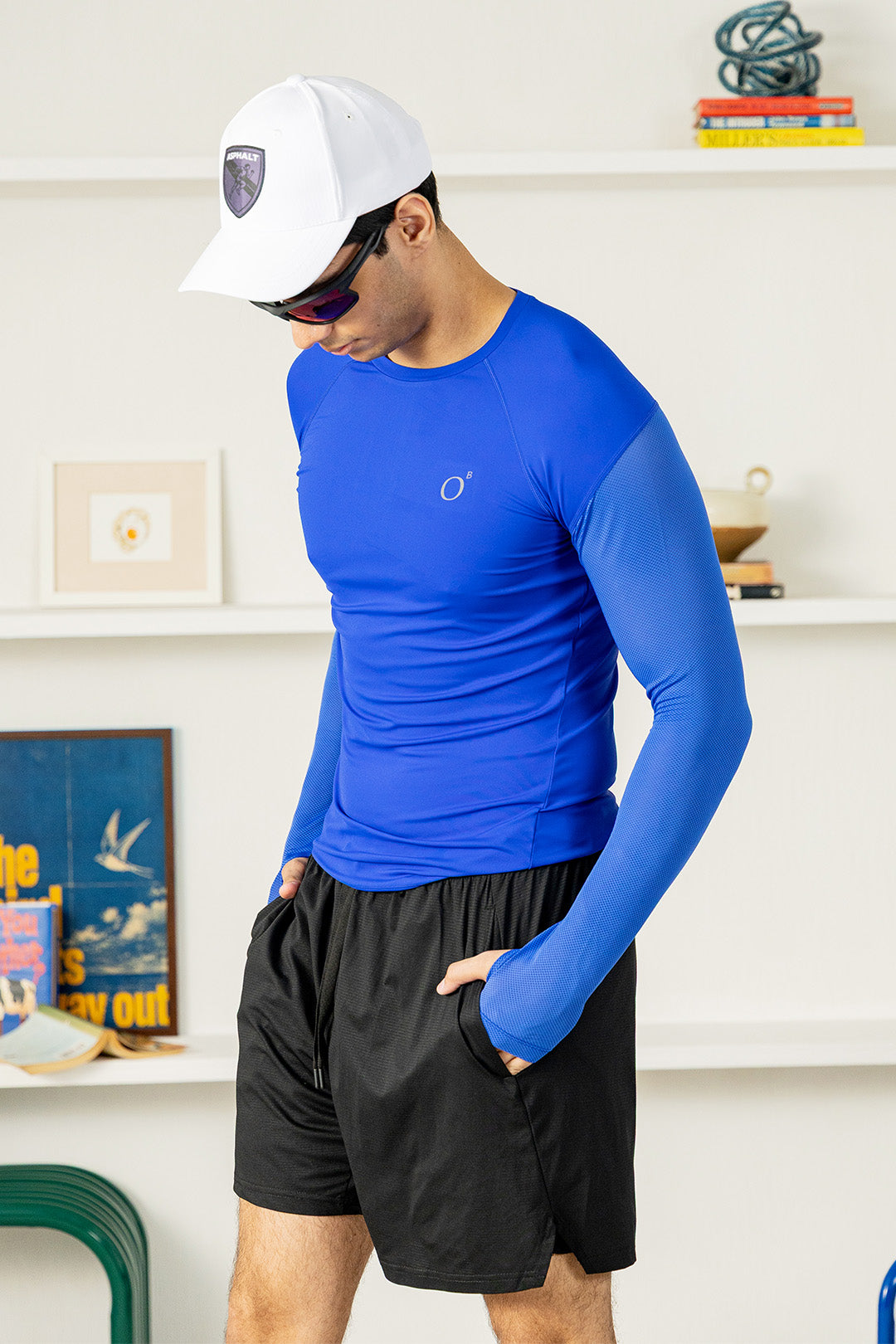 A man wearing a bright blue long-sleeve athletic compression shirt and black athletic shorts, standing in a brightly lit indoor room with white shelves in the background.