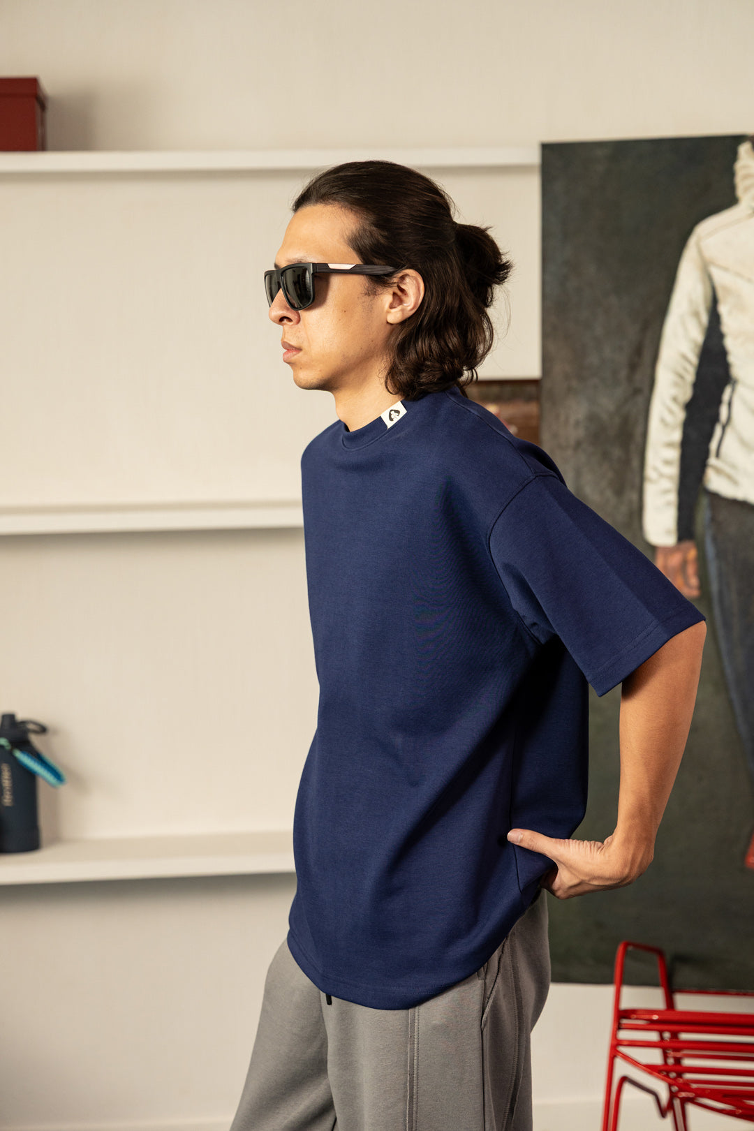 A man wearing a navy blue oversized t-shirt, gray sweatpants, and black sunglasses, standing in a modern indoor setting with white shelves and a framed artwork in the background.