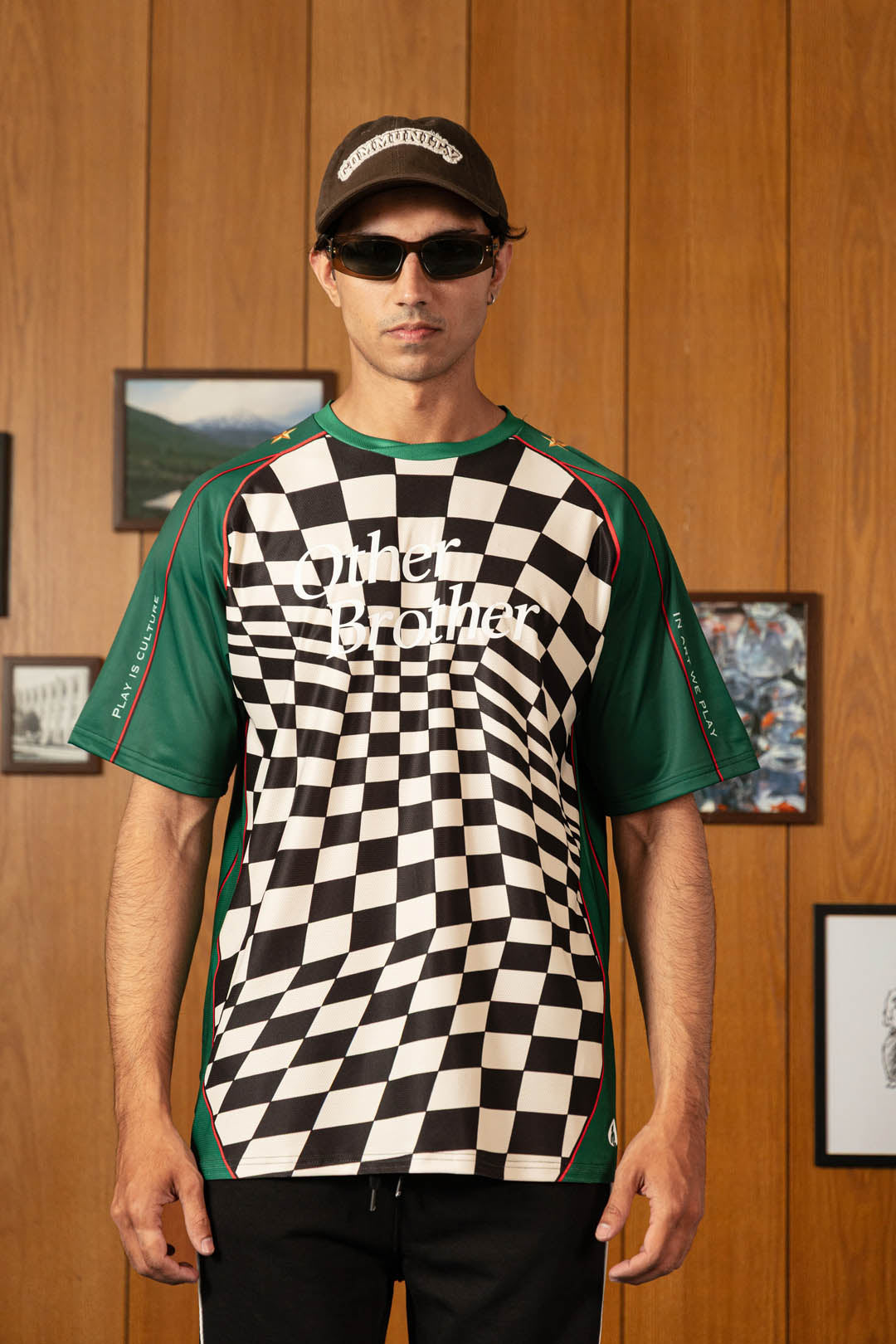 A man wearing a green and black checkered t-shirt with 'Ginet Brother' printed across the chest, paired with a black cap and sunglasses, standing against a wooden paneled wall.