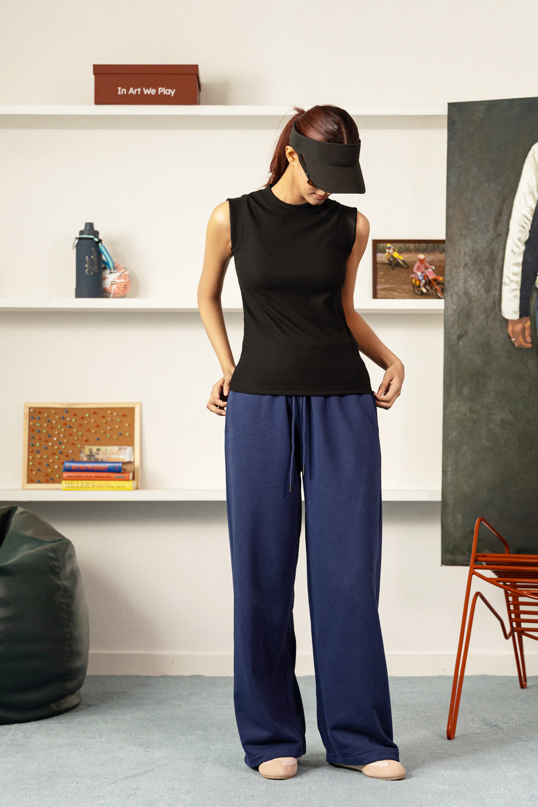 A woman stands in a studio setting wearing a sleeveless black top and navy wide-leg pants, with a black visor over her eyes. Shelves with books and decor are behind her, along with a bean bag and chair.