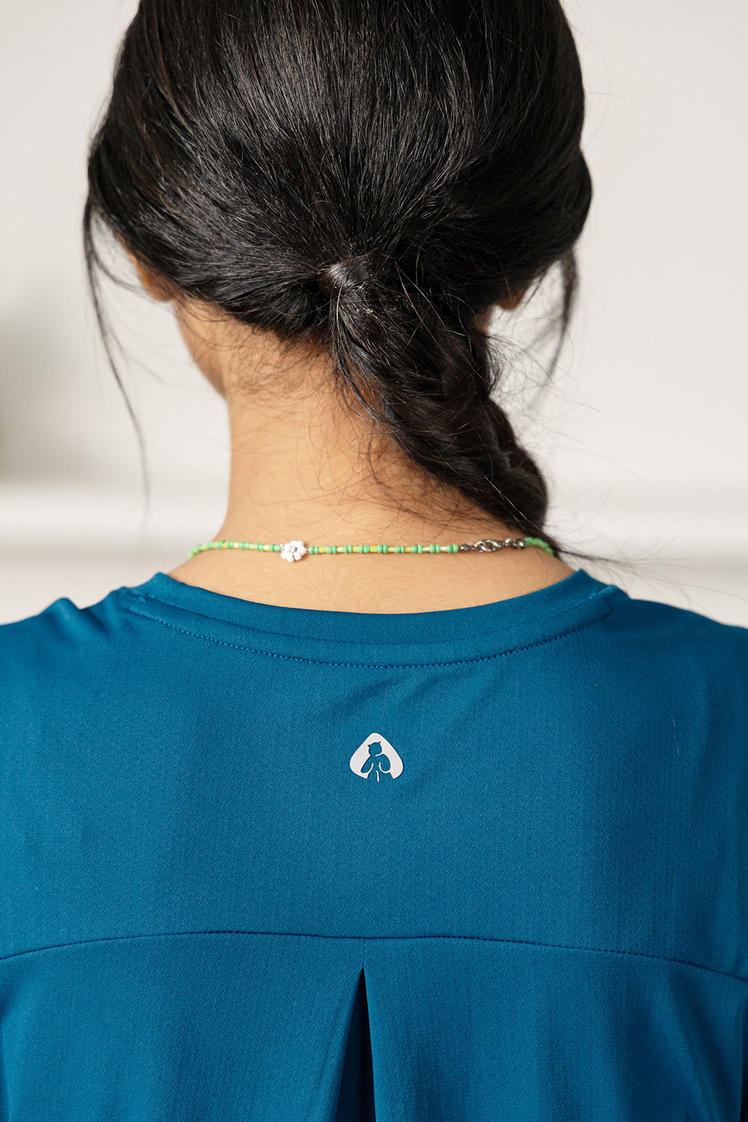 Close-up back view of a person with dark hair in a low bun, wearing a teal athletic top with a small white logo at the center of the back and a thin beaded necklace.