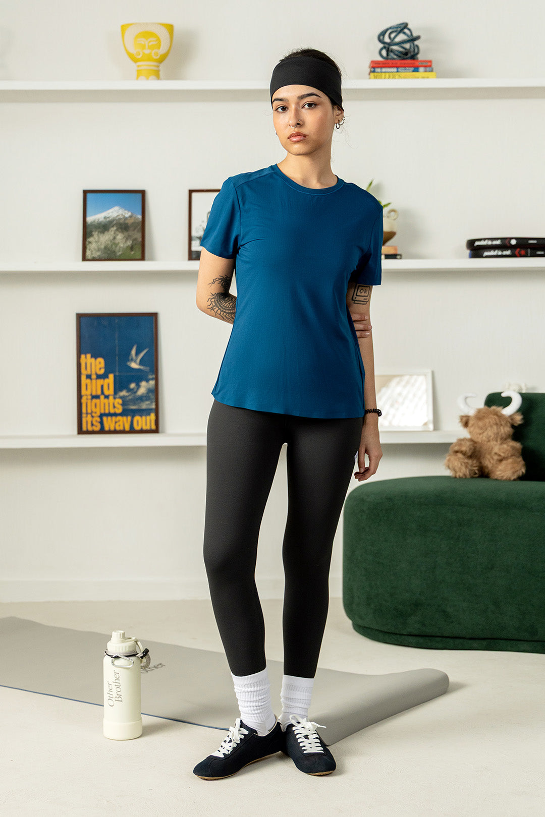 Image of A woman stands in a bright room wearing a fitted short-sleeve athletic T-shirt in deep teal-blue, black leggings, white socks, and dark sneakers. A water bottle, yoga mat, shelving, framed art, and a green ottoman are visible in the background.