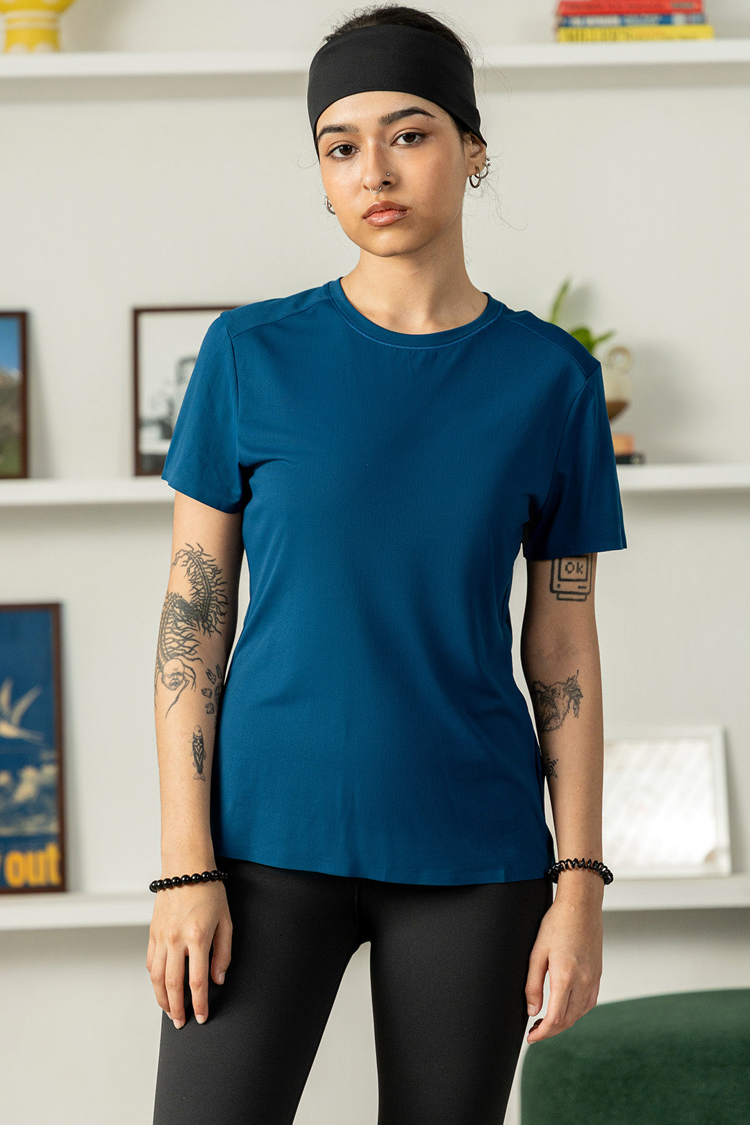 Image of A woman wearing a solid teal blue short-sleeve athletic t-shirt, black leggings, and a black headband, standing in front of a white wall with shelves.