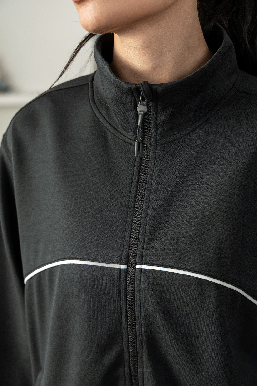 Image of Close-up of a person wearing a black zip-up athletic jacket with a stand collar and a thin white curved stripe across the chest.