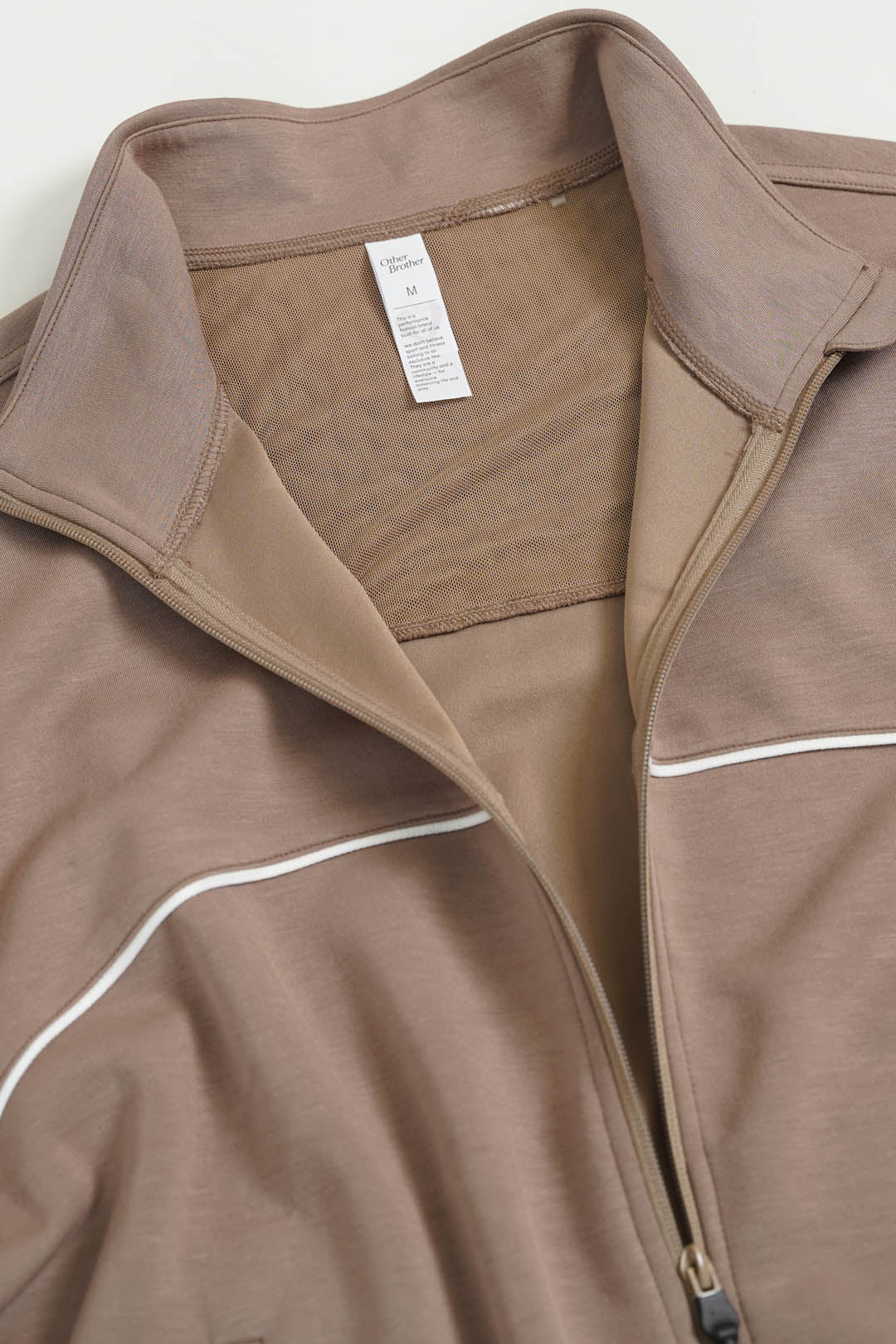 Image of A close-up of a taupe brown zip-up jacket with a stand-up collar and thin white piping details, showing a white interior neck label.