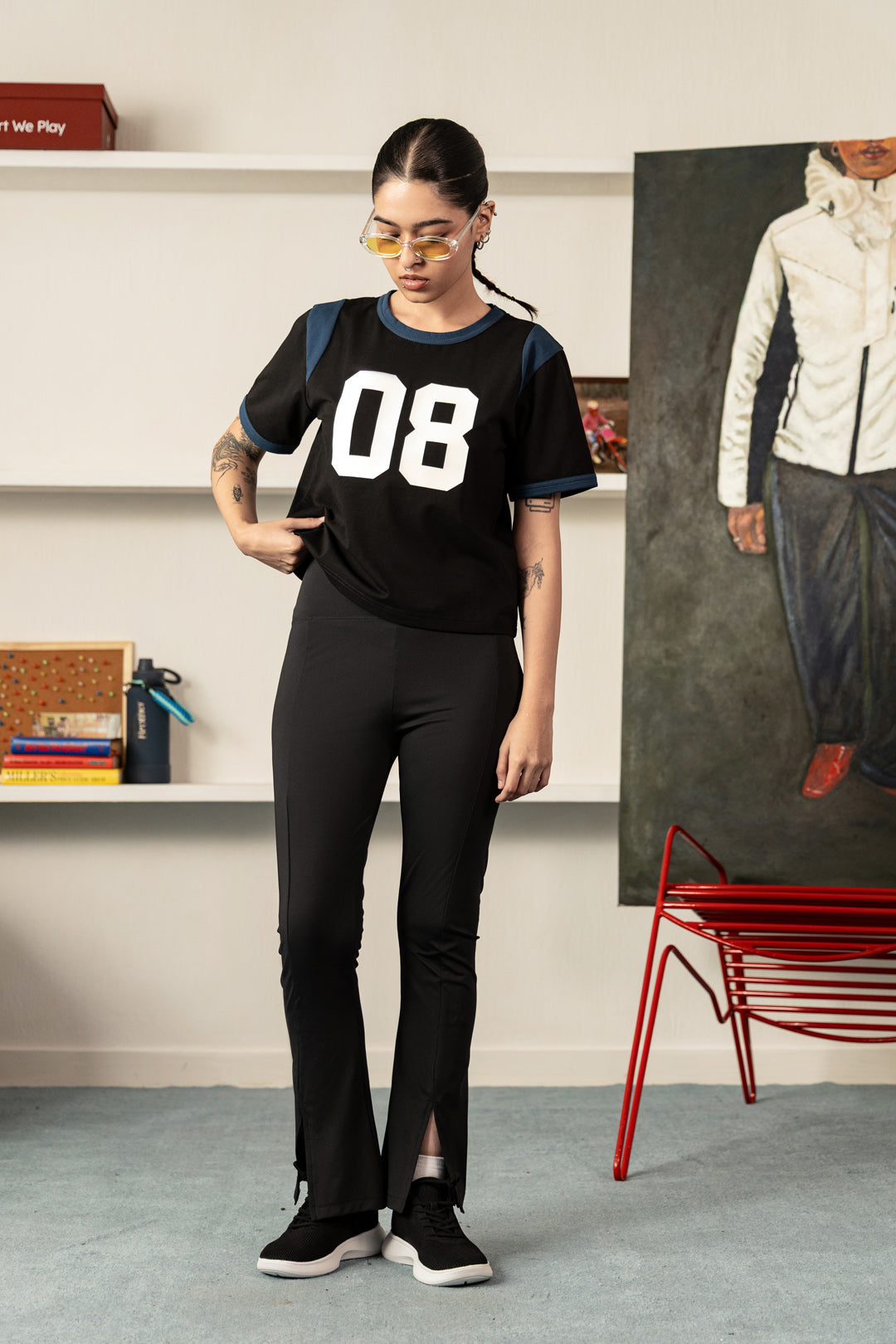 A woman modeling a black short-sleeve t-shirt with '08' printed in white on the front, paired with black pants and black sneakers, standing in a modern room setting.