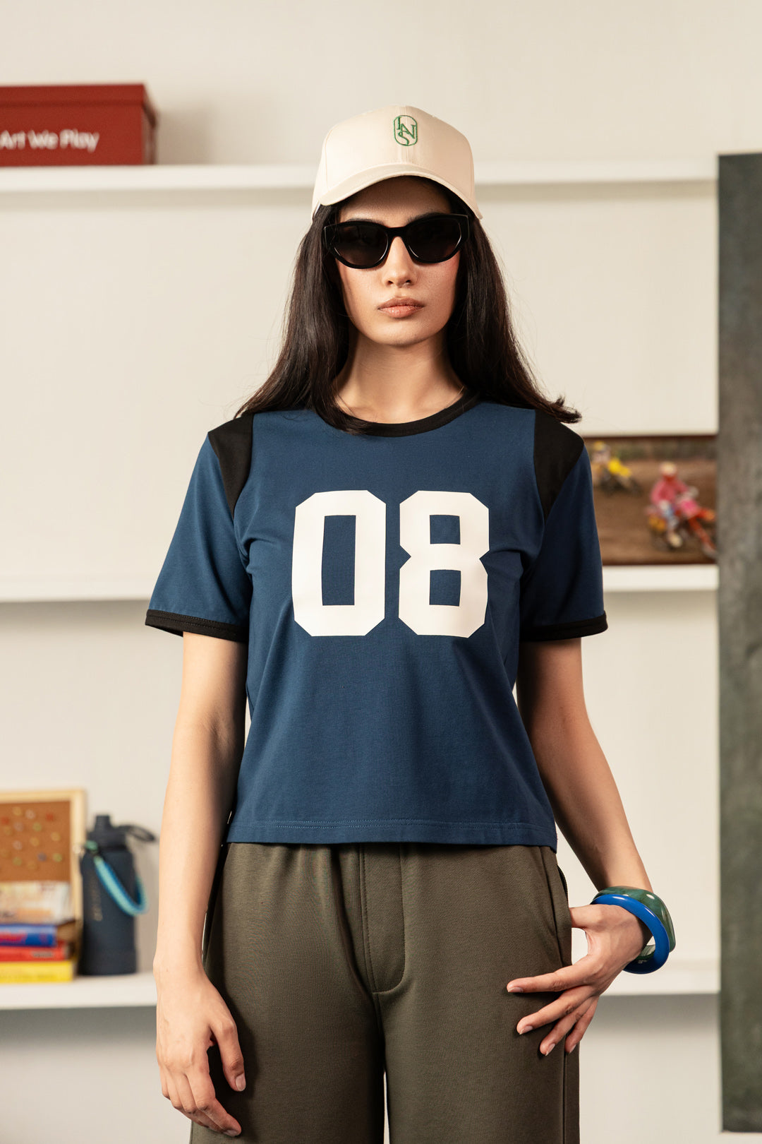 Image of A woman modeling a navy blue short-sleeve t-shirt with black shoulder panels and a large white '08' printed on the front, paired with olive green pants, a beige cap, and black sunglasses.