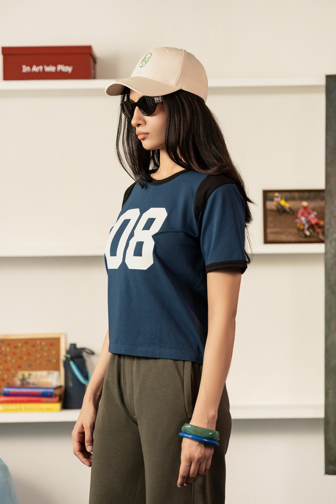 A woman wearing a navy blue cropped t-shirt with black shoulder panels and large white '08' printed on the front, paired with olive green pants, a beige baseball cap, and sunglasses, standing in a room with white shelves.