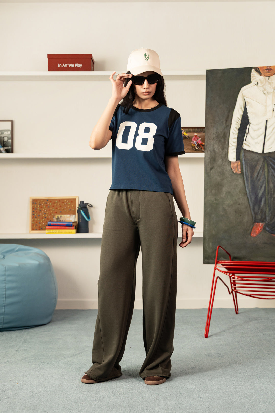 Image of A woman stands in a styled indoor room wearing a navy blue t-shirt with '08' printed in white on the front, olive green wide-leg pants, a beige baseball cap, and black sunglasses.