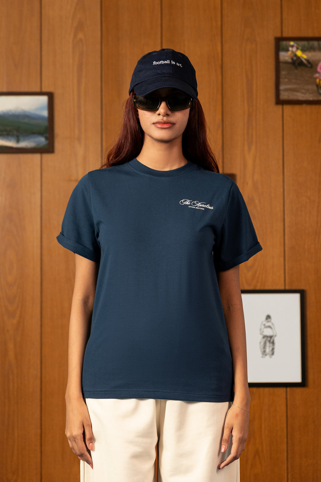 A woman wearing a navy blue crewneck t-shirt with a small white script logo on the left chest, paired with cream pants and a navy baseball cap, standing against a warm wood-paneled wall.