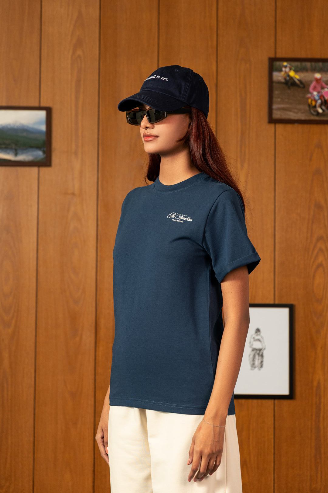 A woman wearing a navy blue short-sleeve t-shirt with a small white script logo on the left chest, paired with cream pants and a navy baseball cap, standing against a wood-paneled wall.