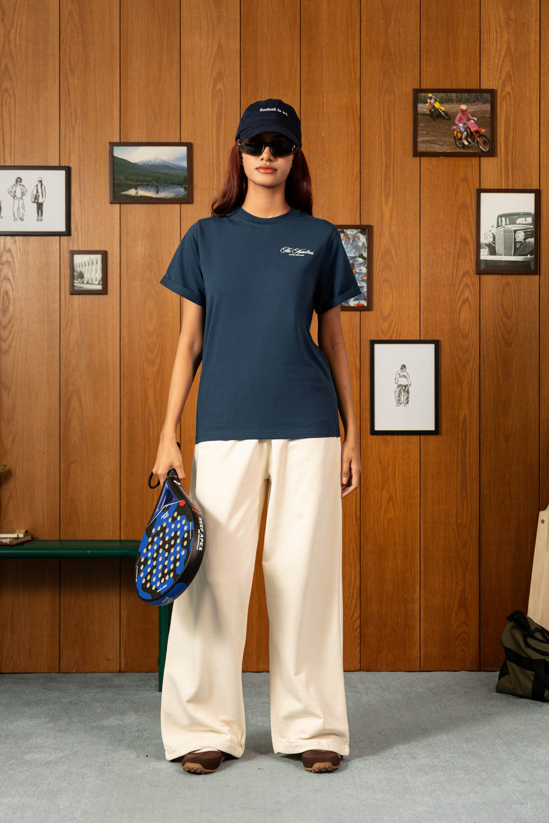 Image of A model stands against a wood-paneled wall wearing a navy blue short-sleeve t-shirt with a small white embroidered logo on the left chest, paired with cream wide-leg pants and a navy baseball cap. She holds a blue and black tennis racket.