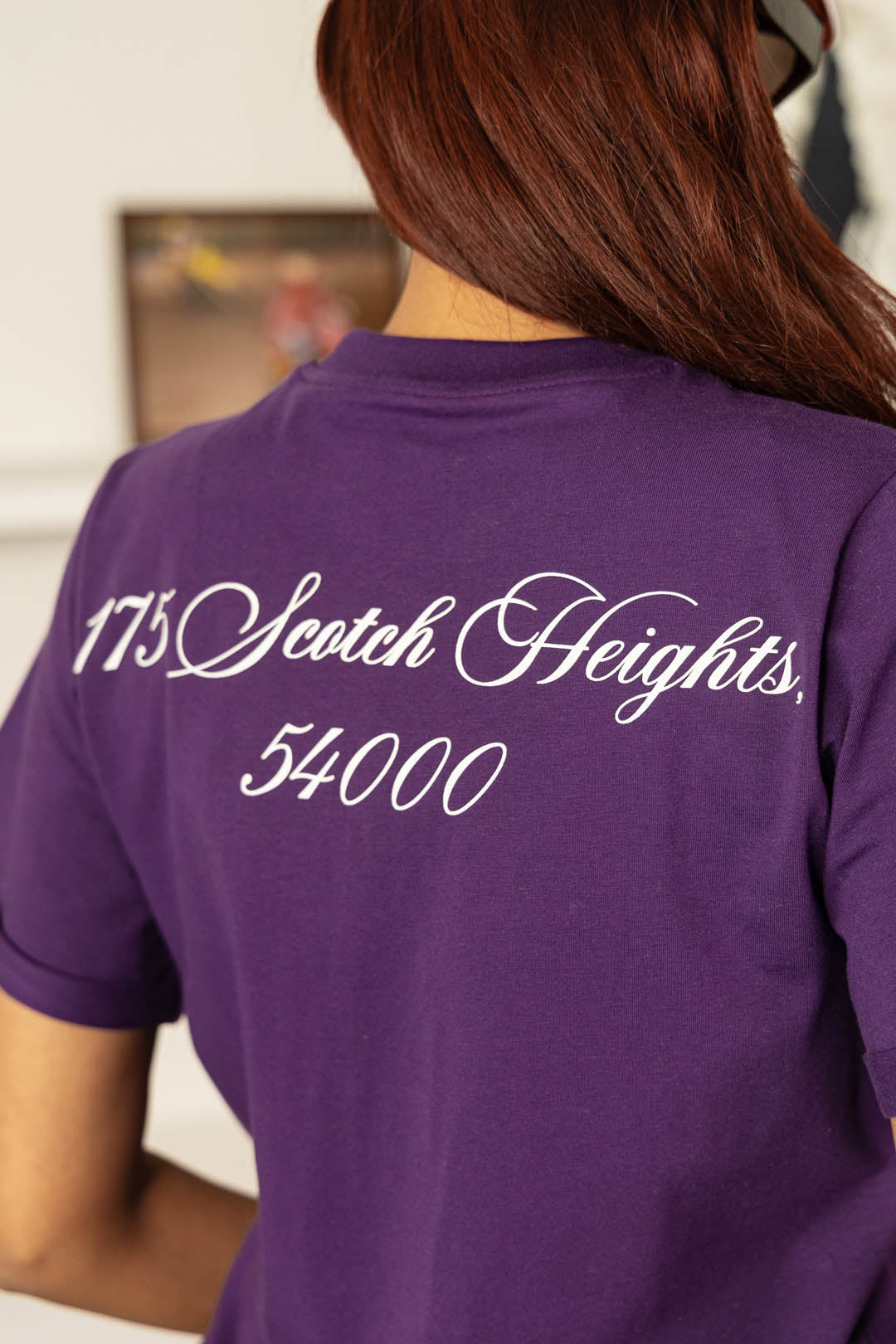 Image of A person shown from the back wearing a purple short-sleeve T-shirt with white script text reading '175 Scotch Heights 54000' across the upper back.