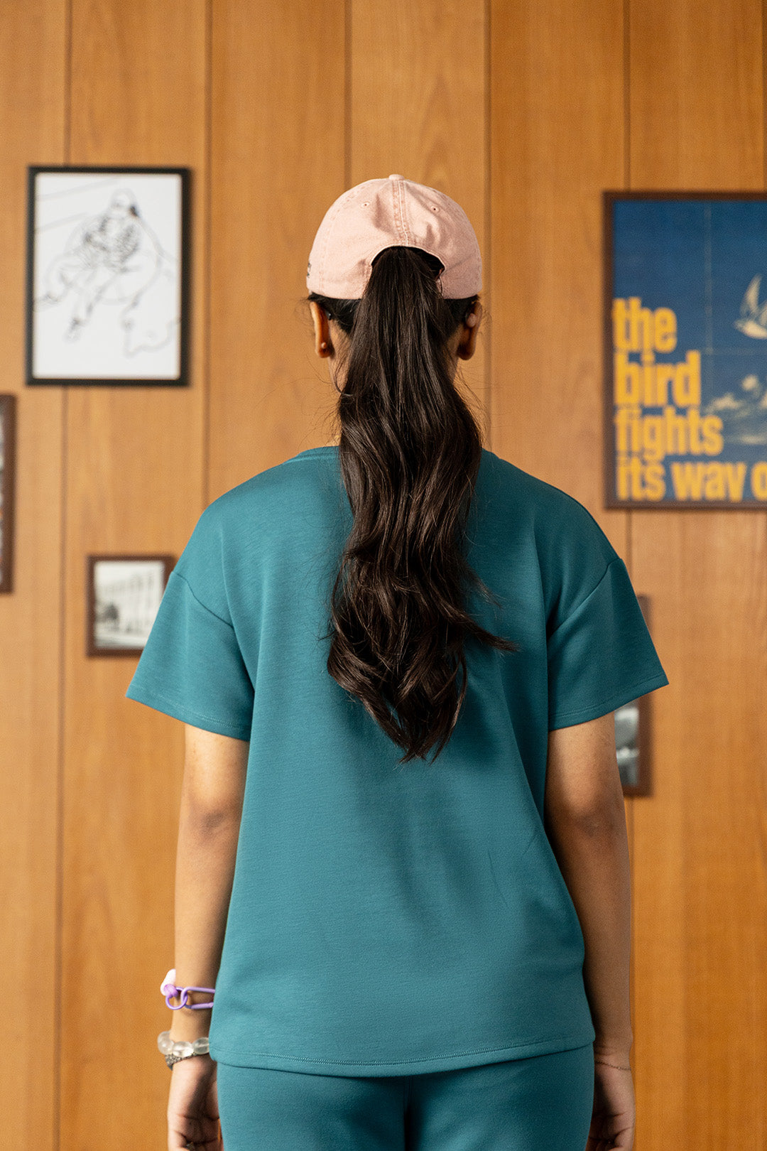 Image of A woman viewed from behind wearing a teal short-sleeve scrub top and matching pants, paired with a light pink baseball cap, standing in front of a wood-paneled wall with framed art.