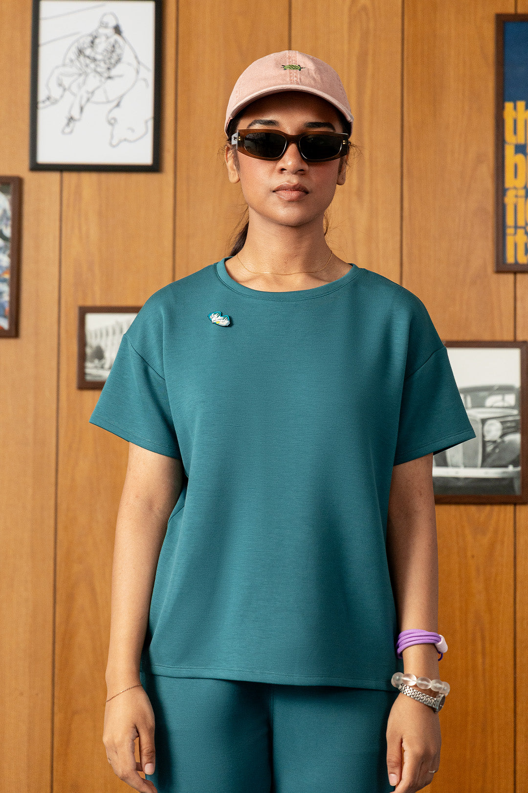 Image of A woman modeling a teal short-sleeve crewneck t-shirt with a small white embroidered logo on the left chest, paired with matching teal leggings, a pink cap, and sunglasses, standing against a wood-paneled wall.