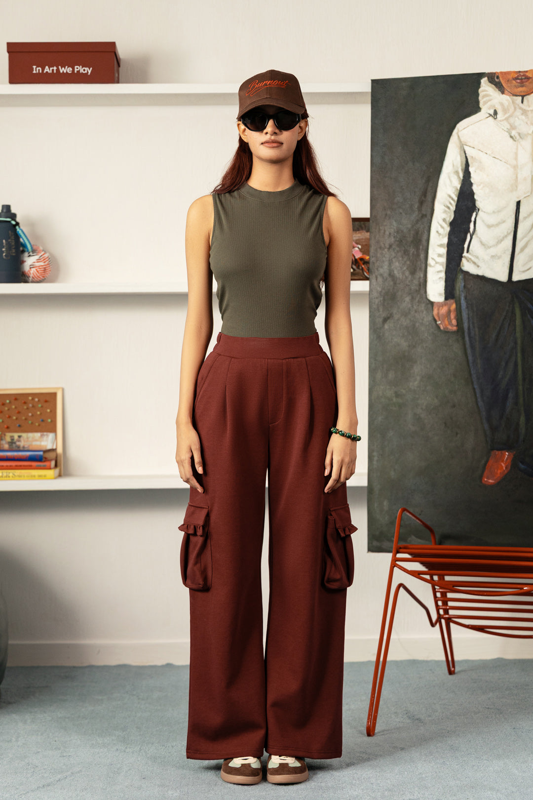 Image of A woman stands in a styled indoor room wearing an olive green sleeveless top, rust-brown wide-leg cargo pants, a brown cap, and sunglasses. The room has white shelves with decor, a large portrait painting, and a red chair.