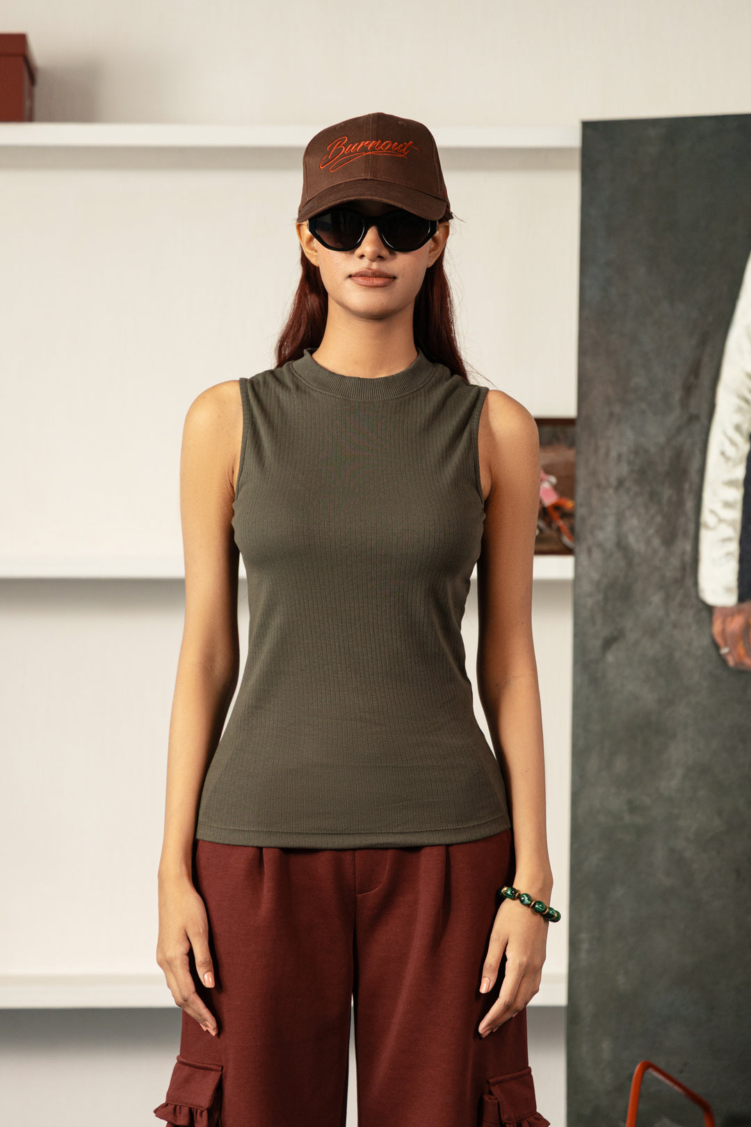 A woman modeling a sleeveless ribbed knit top in olive green, paired with rust-brown cargo pants, a brown cap, and black sunglasses, standing in an indoor studio setting.
