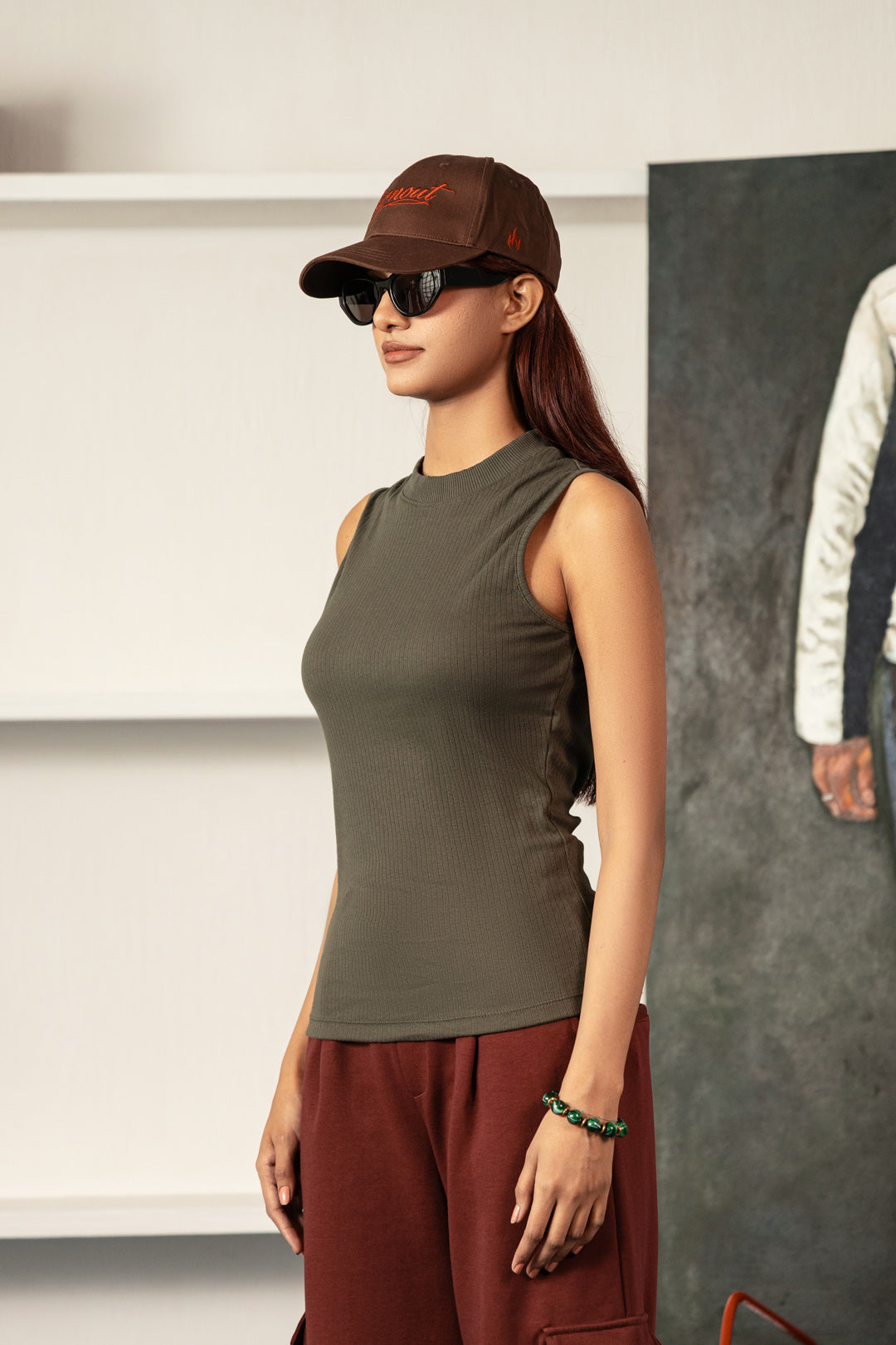Image of A woman wearing a ribbed sleeveless crop top in olive green, styled with a brown cap, sunglasses, and rust-colored pants, standing in a modern indoor setting.