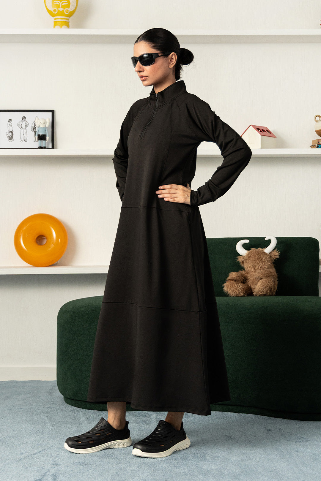 A woman modeling a long-sleeved black maxi dress with a mock neck and side pockets, styled with black sneakers and sunglasses in a modern living room setting.