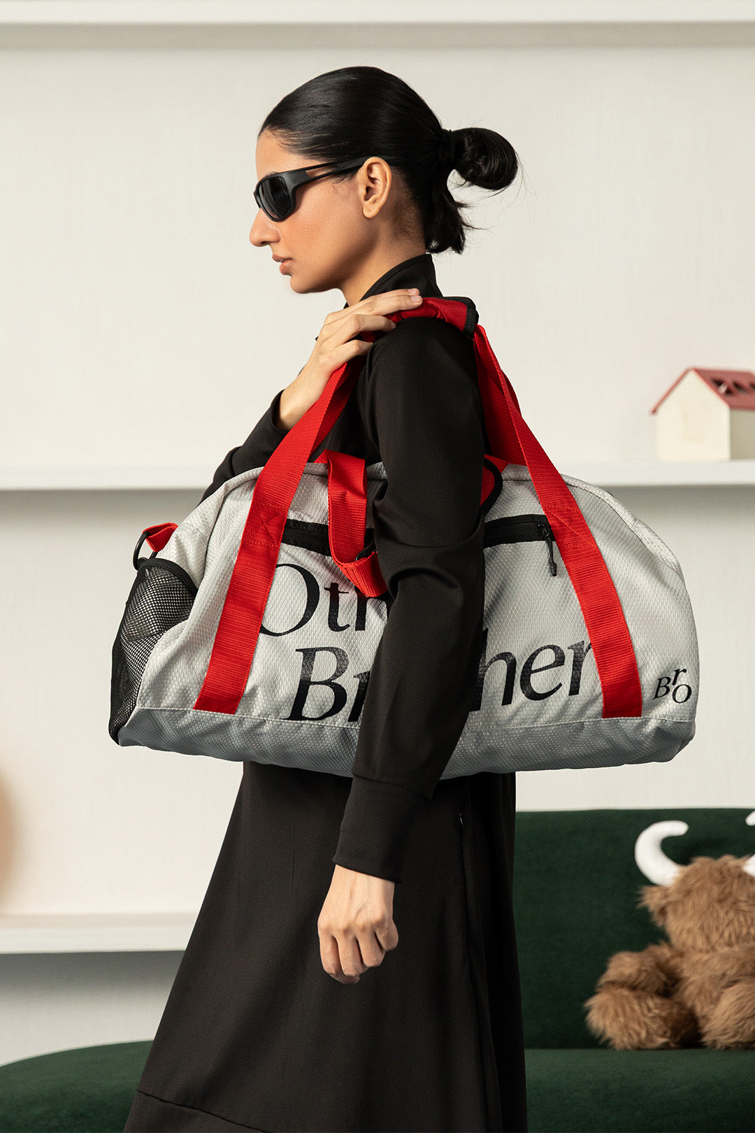 Image of A woman in a black outfit and sunglasses carries a large gray duffel bag with bold red shoulder straps and black lettering, set against a modern indoor background.