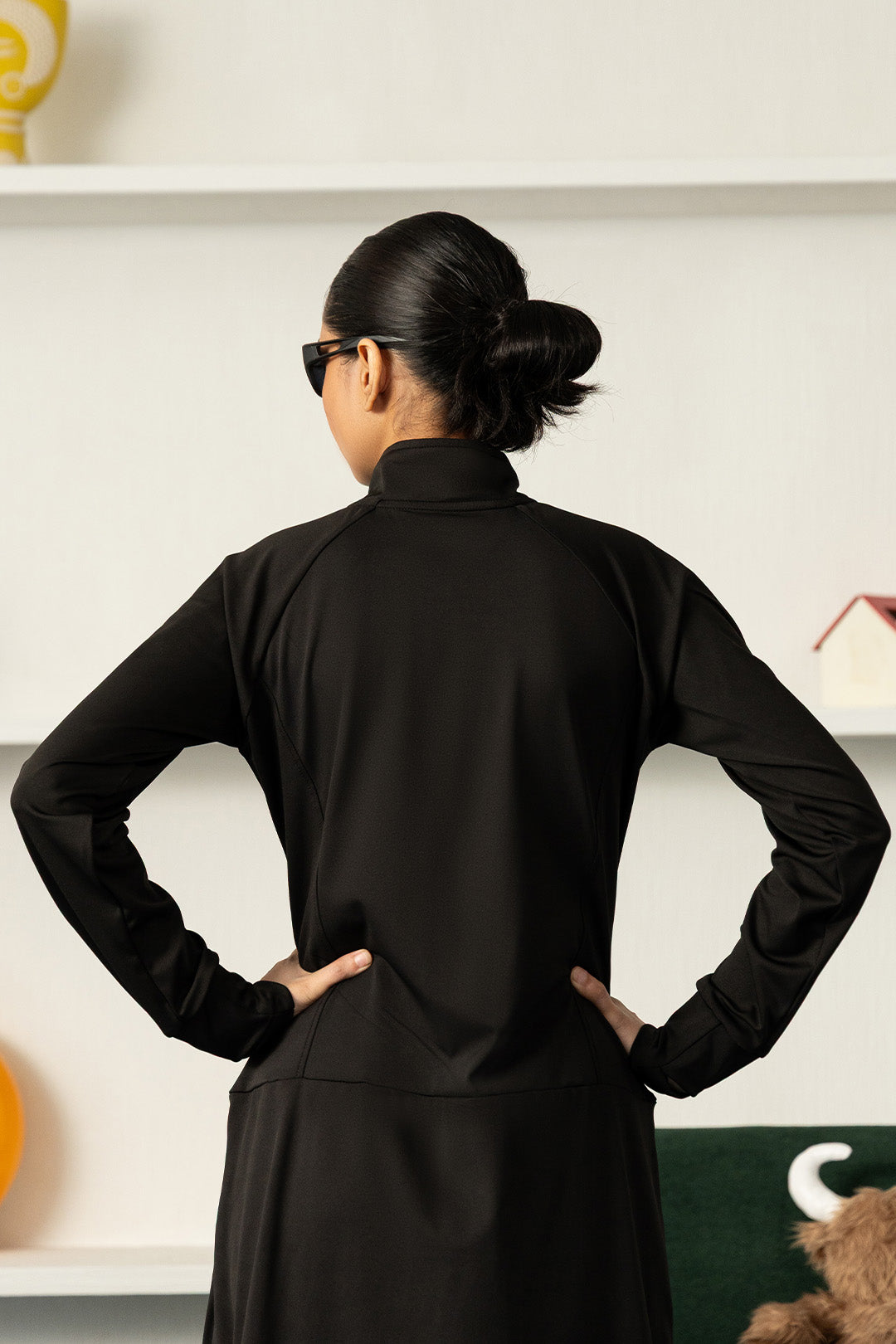 A woman viewed from behind wearing a black long-sleeve mock-neck athletic top and matching black bottoms, standing with hands on hips in an indoor setting.