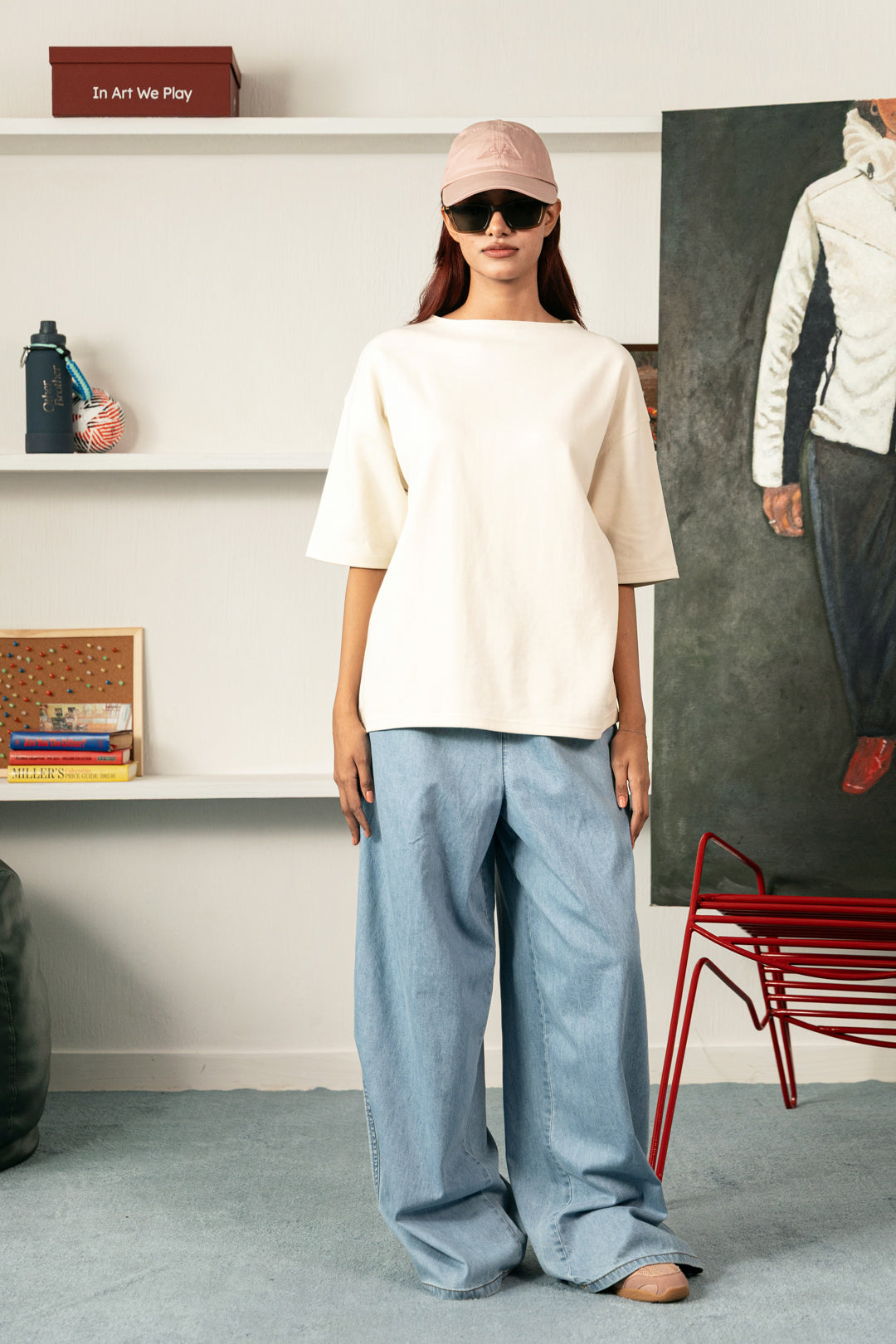Image of A model stands in a styled room wearing an oversized cream-colored short-sleeve top and wide-leg light blue jeans, paired with a pink cap and sunglasses.