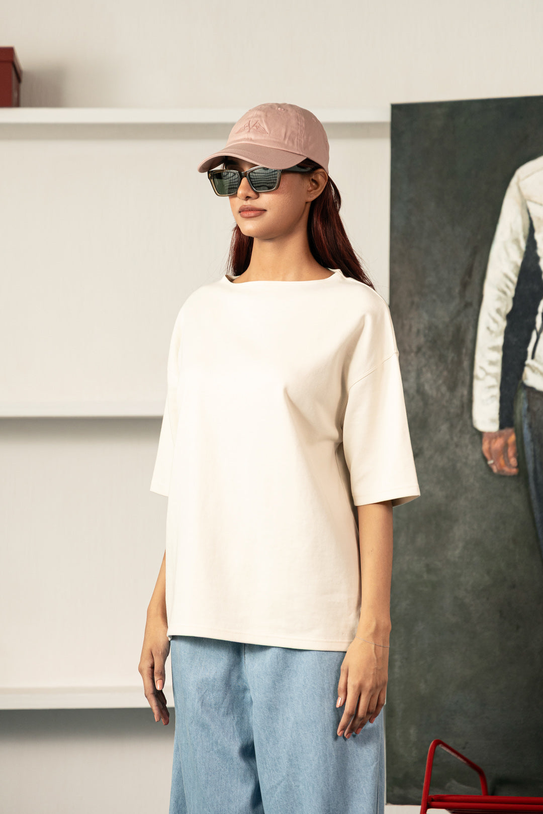 Image of A woman wearing an oversized off-white short-sleeve t-shirt, light blue wide-leg jeans, a dusty pink baseball cap, and black sunglasses, standing in a modern indoor setting.