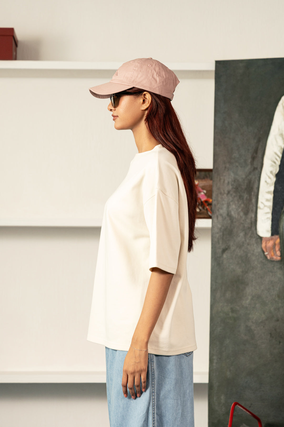 A woman in profile wearing an oversized off-white t-shirt, light blue jeans, a dusty pink baseball cap, and sunglasses, standing in a modern indoor setting with white shelves and a dark textured panel.