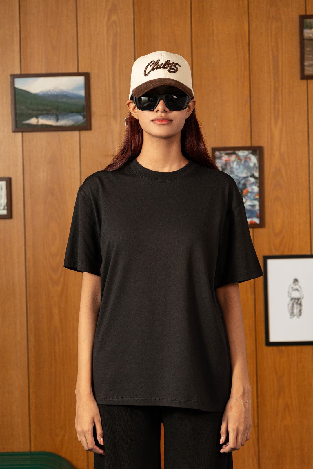 Image of A woman stands against a wood-paneled wall wearing an oversized black short-sleeve crewneck T-shirt, black pants, a cream baseball cap with brown lettering, and black sunglasses.
