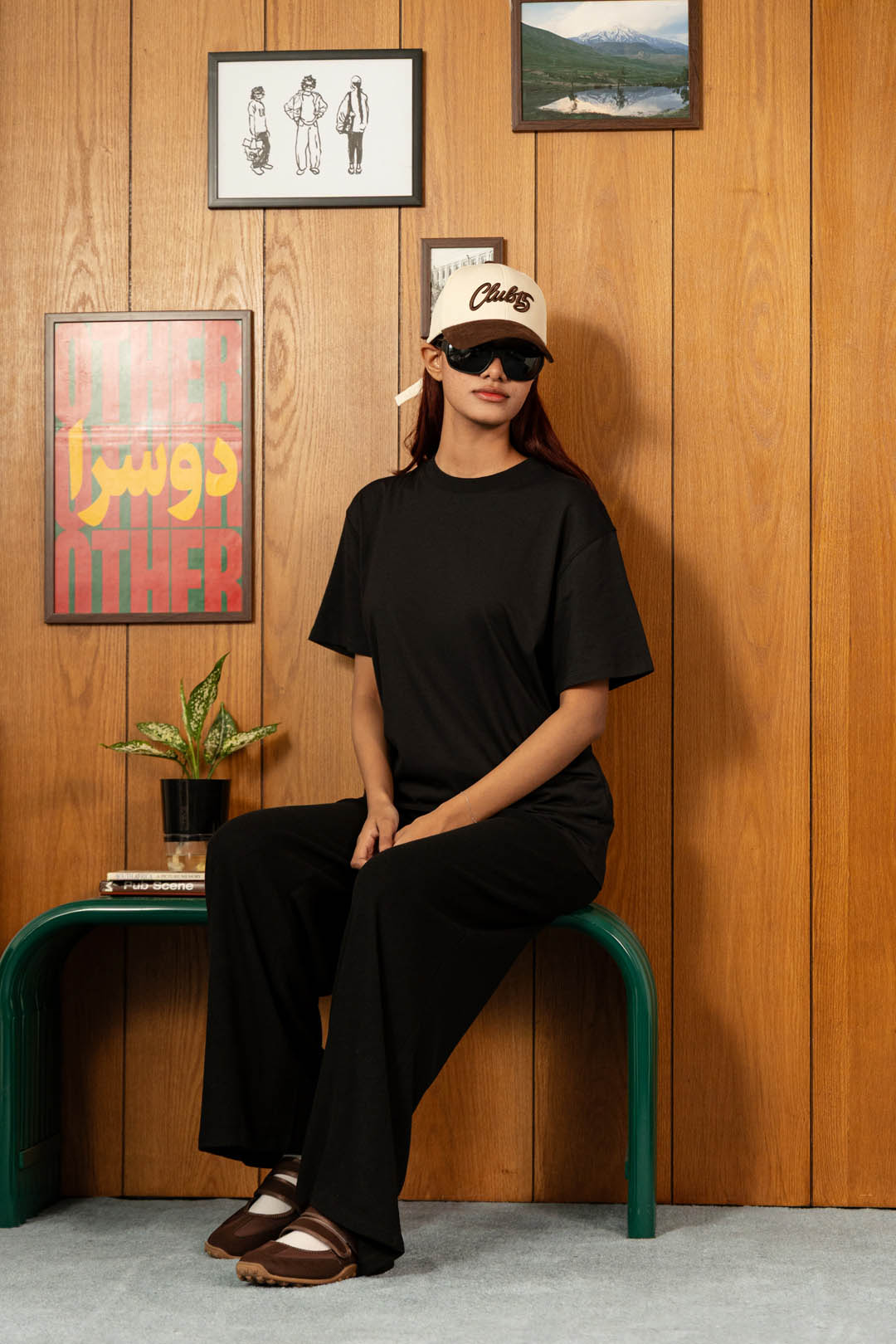A woman wearing a black oversized t-shirt and black wide-leg pants sits on a green bench against a wood-paneled wall. She is wearing a beige baseball cap with 'Chilewich' written on it and dark sunglasses.
