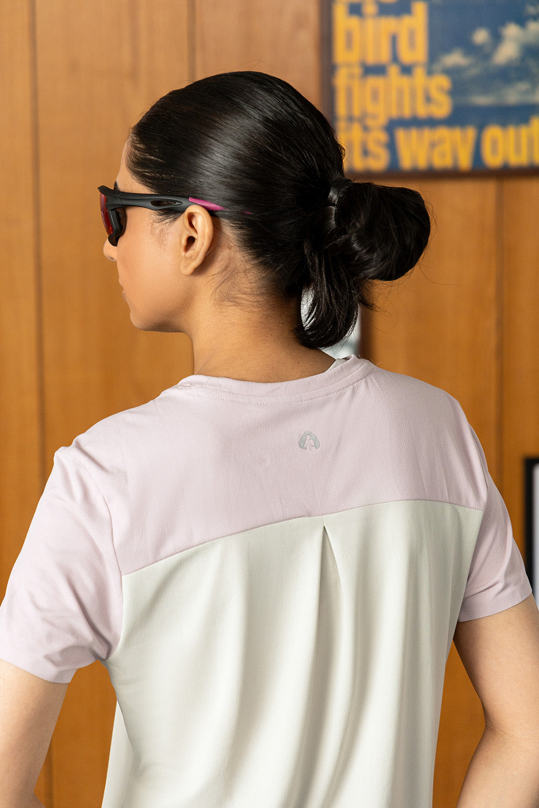 Image of A woman shown from the back wearing a color-blocked athletic t-shirt with a light pink upper section and an off-white lower section, paired with black sunglasses.