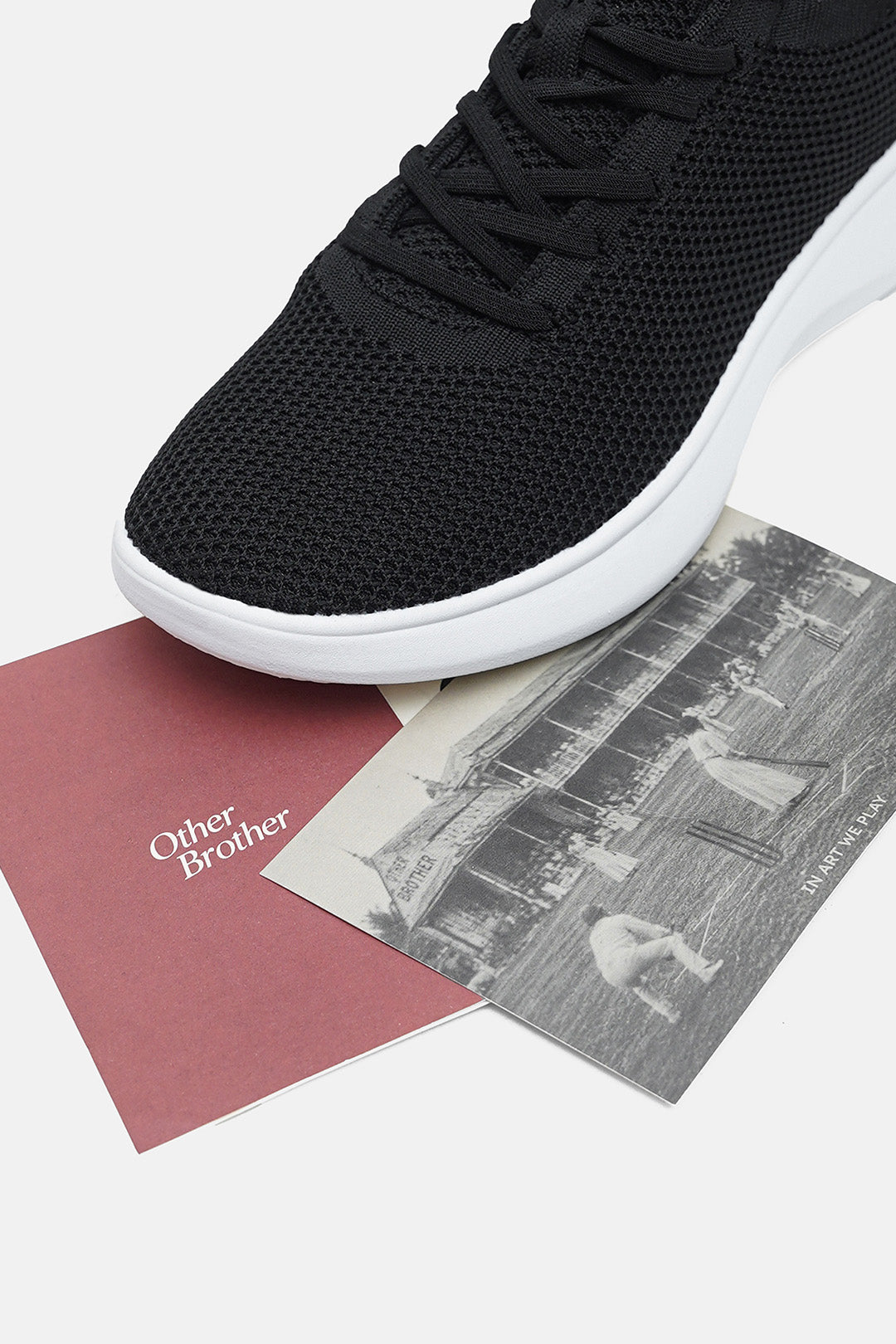 A close-up of a black knit sneaker with a white sole, resting on a dusty rose card labeled 'Other Brother' and a vintage black-and-white photograph, on a light gray background.